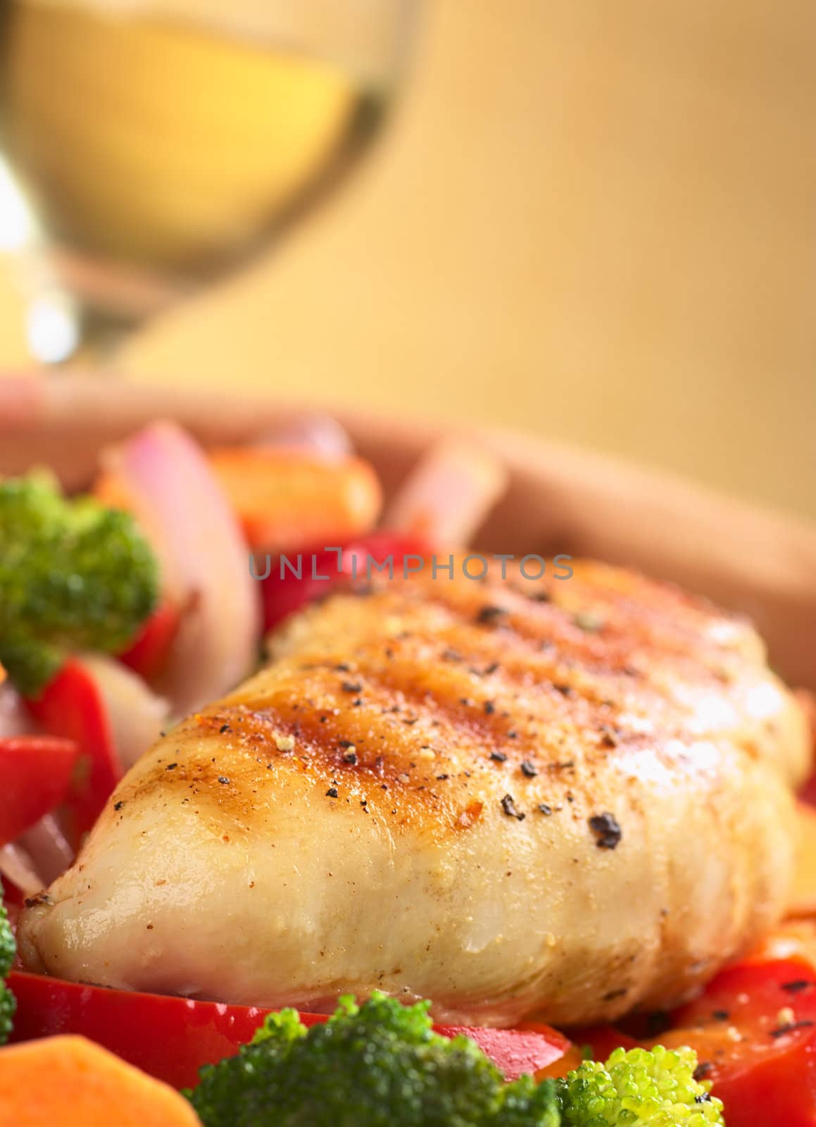 Fried chicken breast with pepper on top surrounded by vegetables (onion, carrot, broccoli, red bell pepper) with white wine in the background (Selective Focus, Focus on the front of the meat)