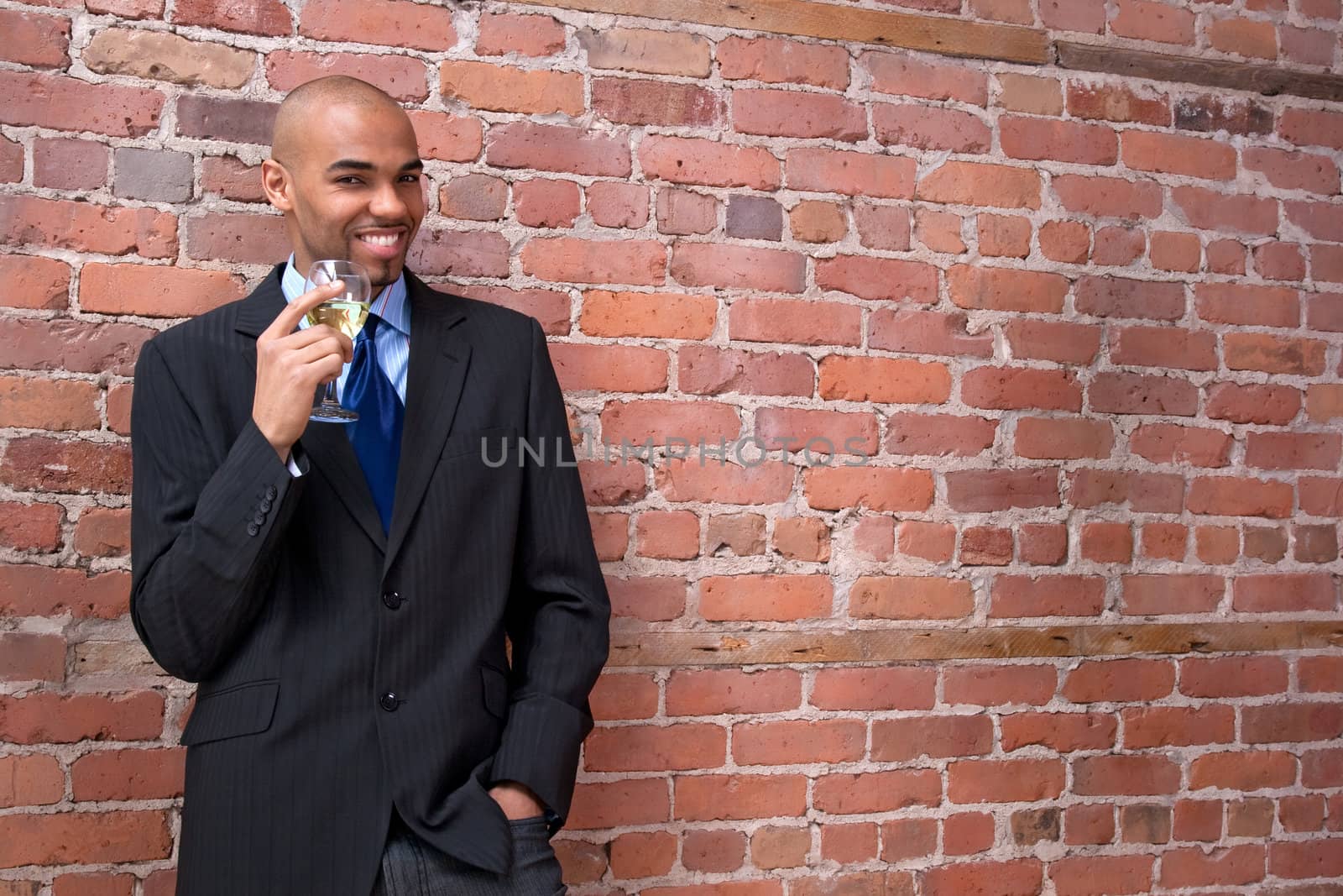 Young business man leaning against the brick wall, drinking wine and smiling.