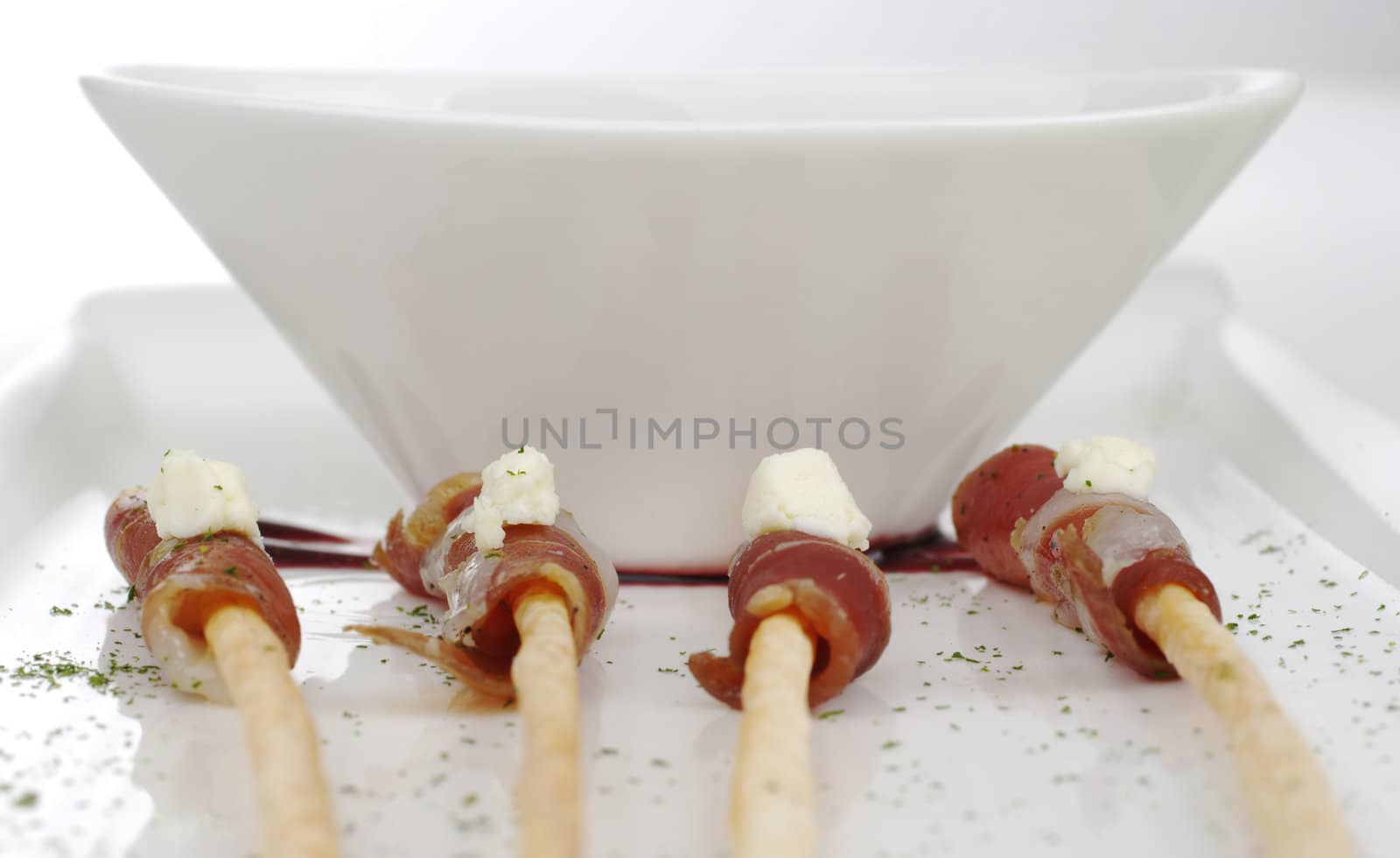 Thin ham slices wrapped around thin bread sticks and served on a long plate with a bowl in the background (Selective Focus) 