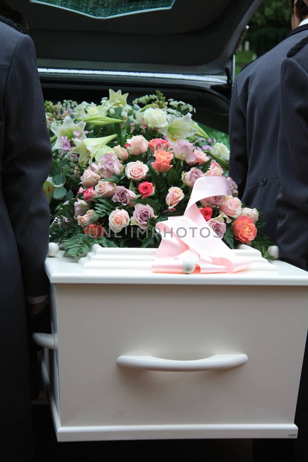 A white coffin, covered with flowers near a hearse, people taking out the coffin