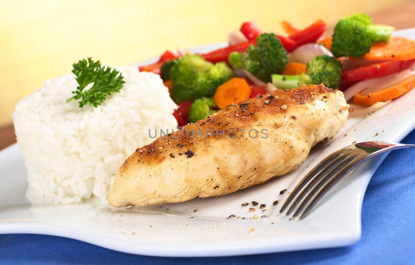 Chicken breast with pepper on top, vegetables (carrot, broccoli, onion, red bell pepper) and rice garnished with parsley (Selective Focus, Focus on the front of the meat)