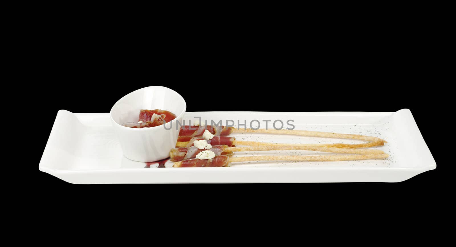 Thin bacon slices wrapped around salt sticks and served on a long plate with a bowl containing bacon isolated on black photographed from the side (Selective Focus)  