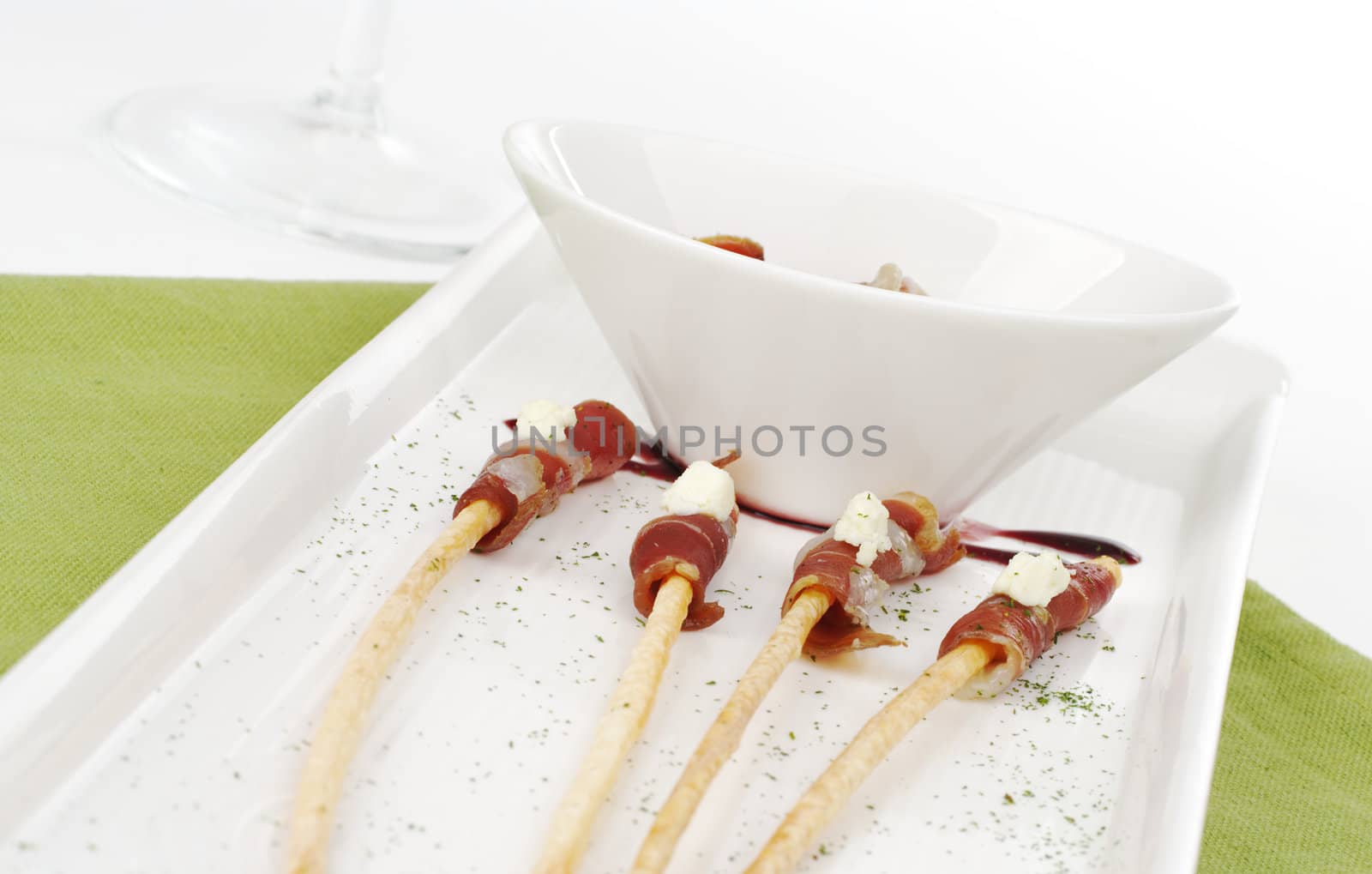 Thin ham slices wrapped around thin bread sticks and served on a long plate on a green place mat with a wine glass in the background (Selective Focus) 