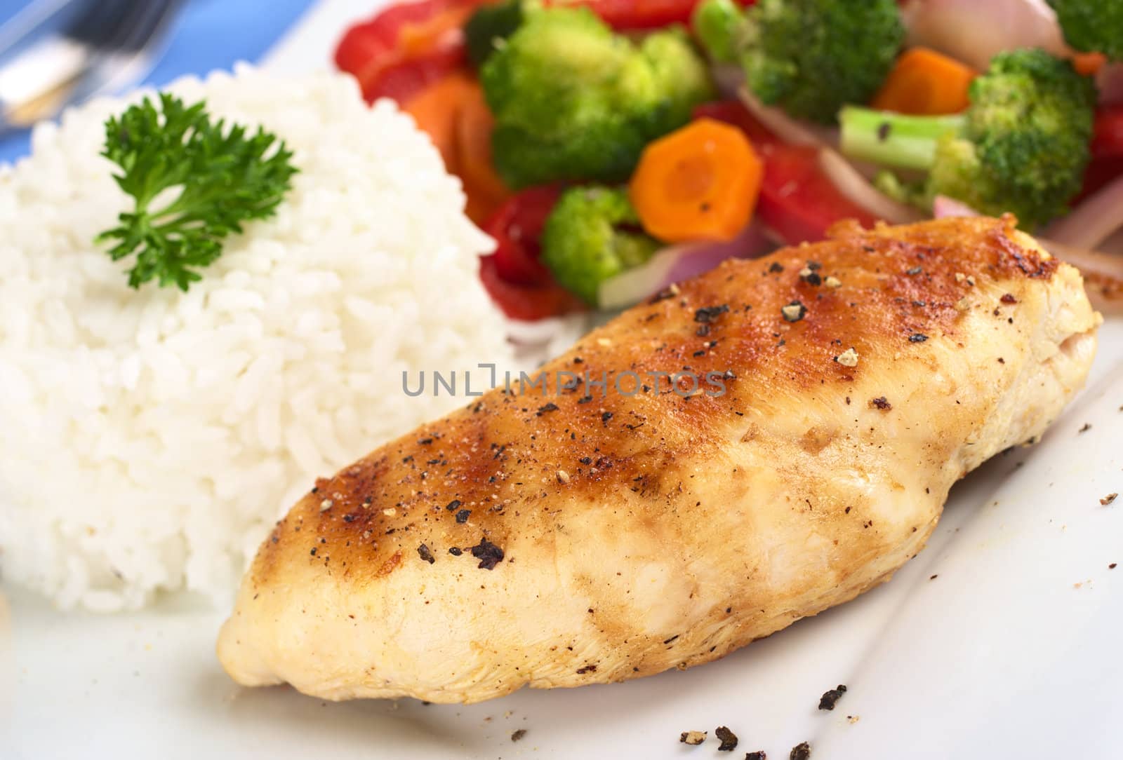 Fried chicken breast with pepper on top, vegetables (carrot, broccoli, onion, red bell pepper) and rice garnished with parsley (Selective Focus, Focus on the front of the meat)