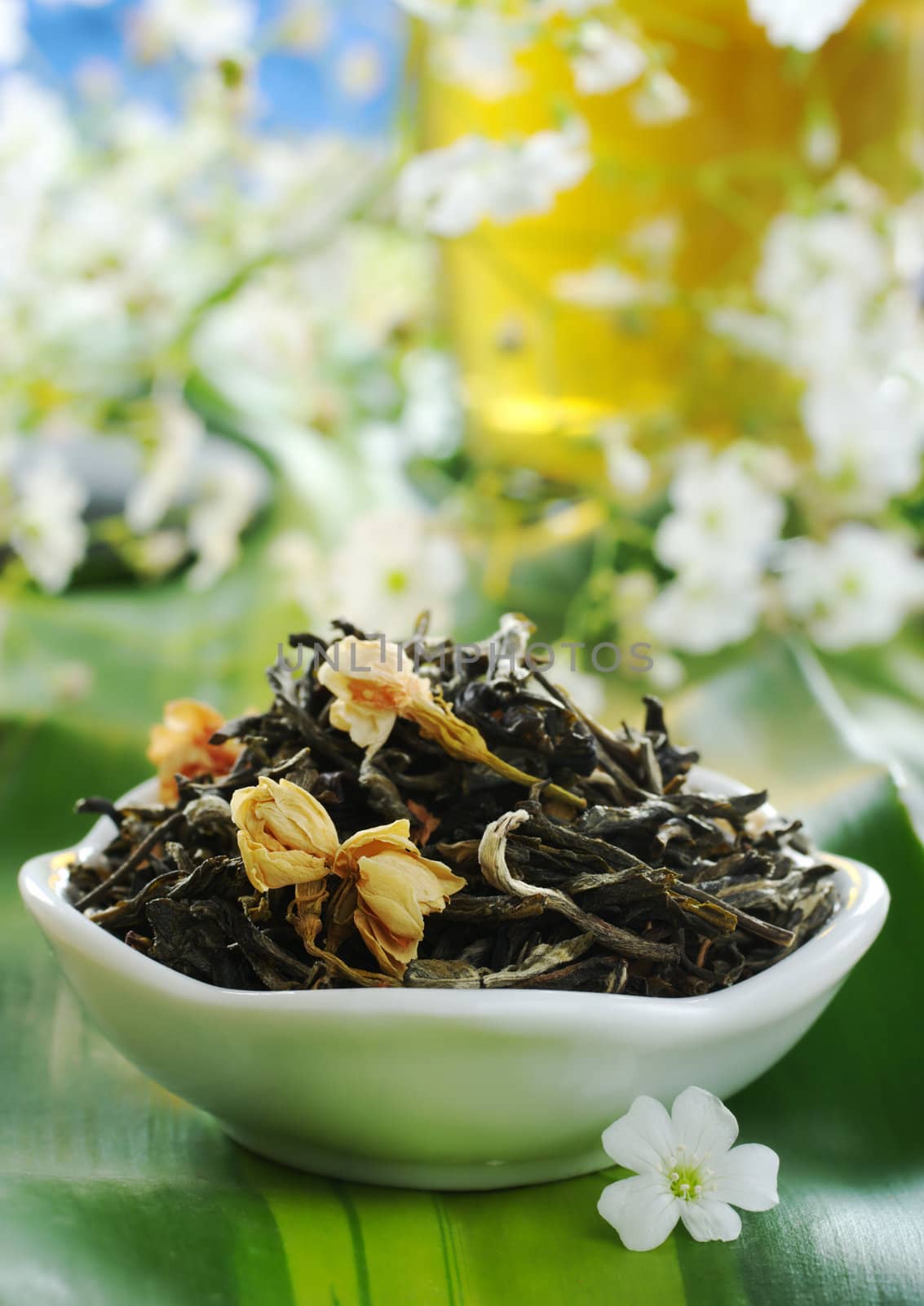 Loose green tea with jasmine flowers in a bowl with a glass cup of tea and some white flowers in the background (Selective Focus, Focus on the front of the loose tea and the jasmine flowers) 