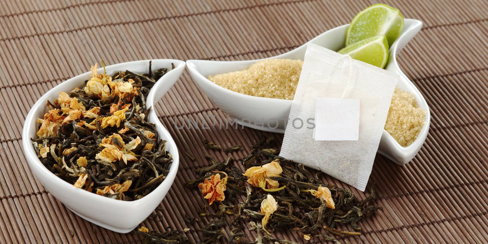 Teabag with an empty label, green tea with jasmine flowers, and brown sugar with lemon pieces in bowl on brown table mat (Selective Focus, Focus on the teabag)