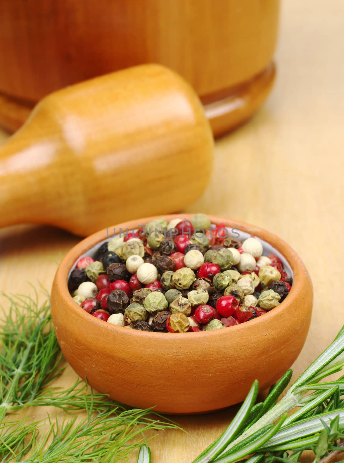 Black, white, green and red pepper corns in a ceramic bowl with other herbs (rosemary, dill) in the foreground and a mortar and pestle in the background (Selective Focus, Focus on the front of the bowl and the pepper corns) 