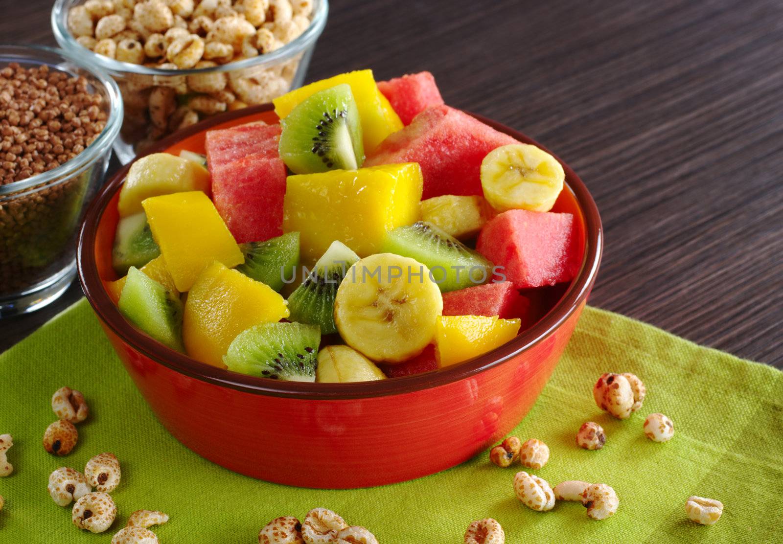 Fresh fruit salad made of banana, kiwi, watermelon and mango pieces in orange bowl with cereals (puffed wheat and puffed chocolate quinoa) (Selective Focus, Focus on the front of the bowl and the fruits in the front)