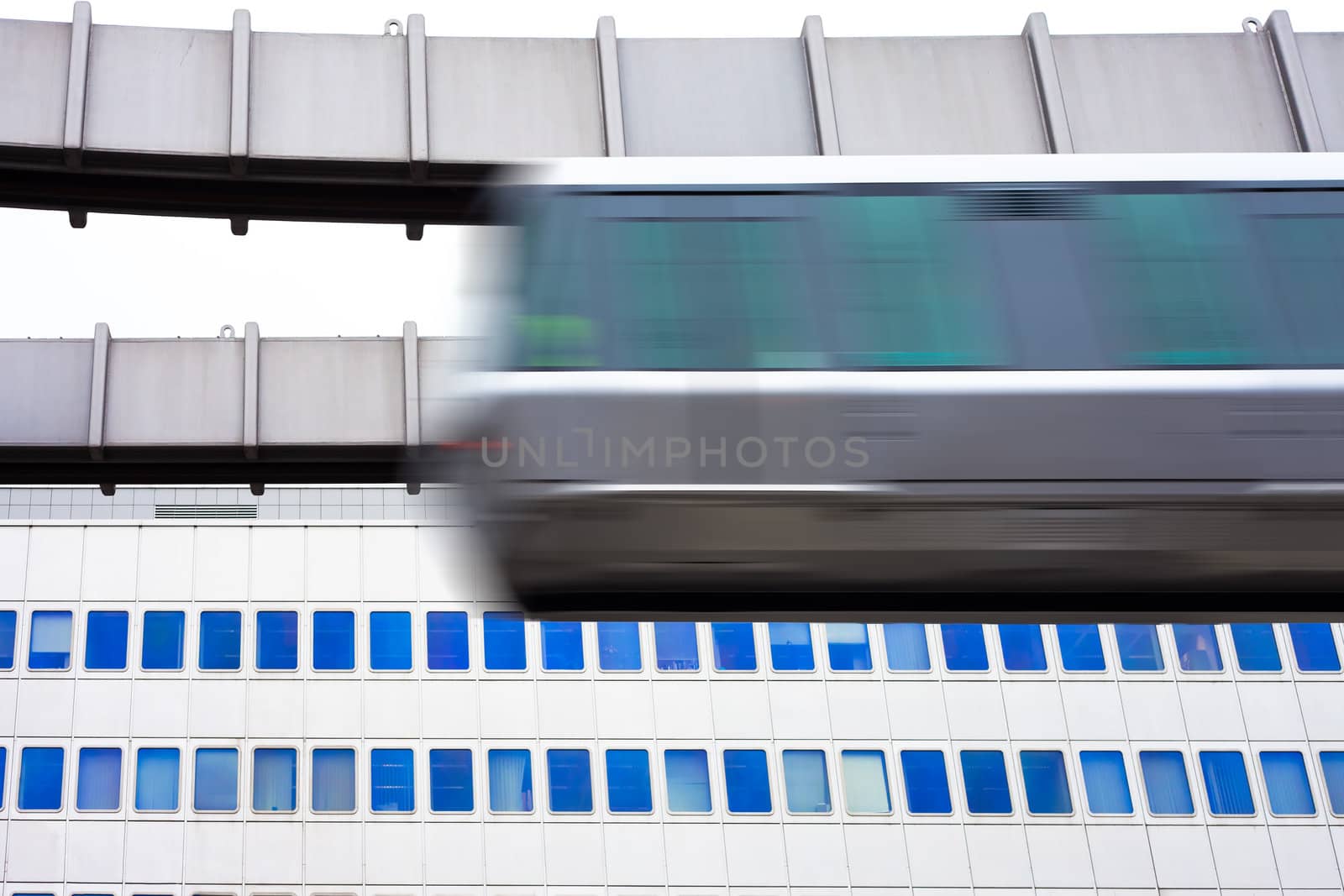 Modern public transportation system skytrain hanging from elevated guideway passes by in front of tall office building facade (with motion blur).