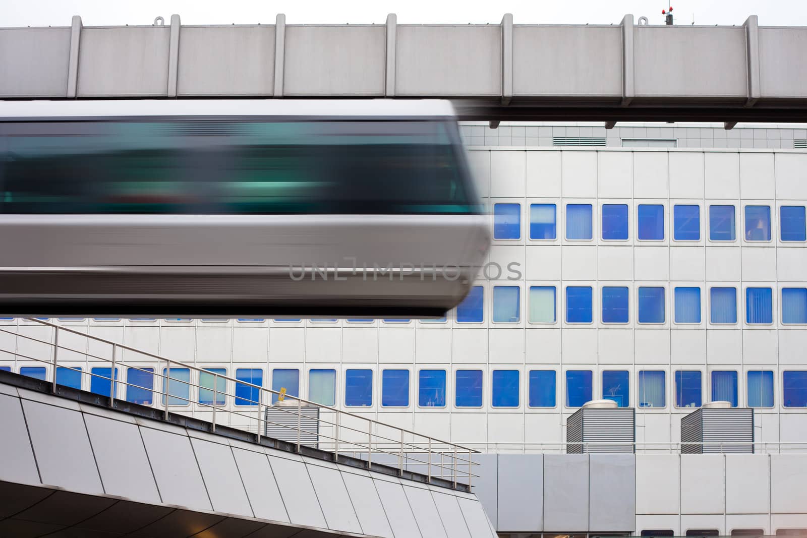 Modern public transportation system skytrain hanging from elevated guideway passes by in front of tall office building facade (with motion blur).