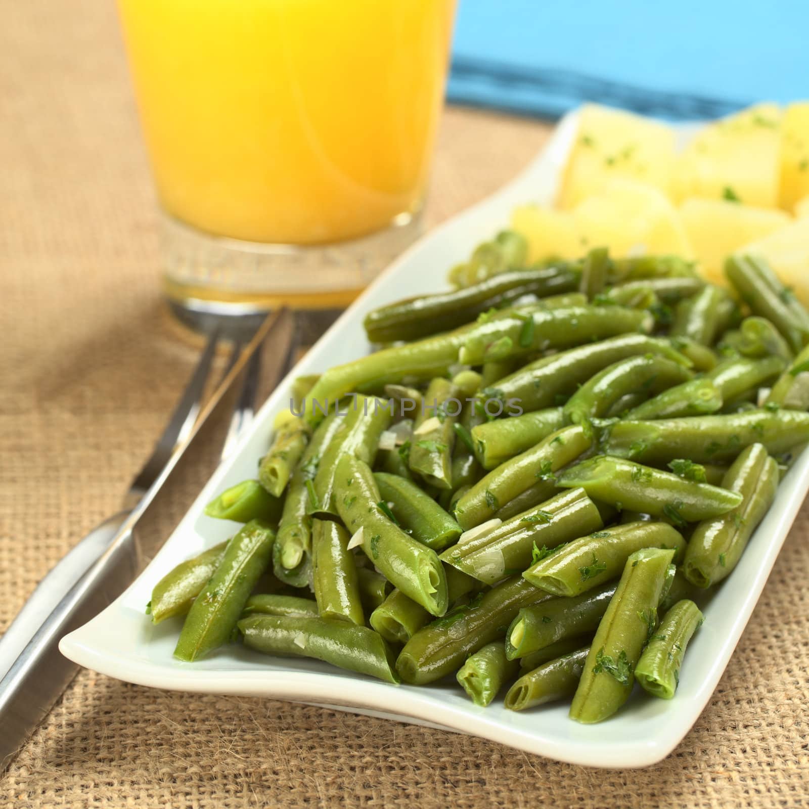 Cooked green beans with onion and parsley with cooked potato, orange juice and blue napkins in the back (Selective Focus, Focus on one third of the picture)