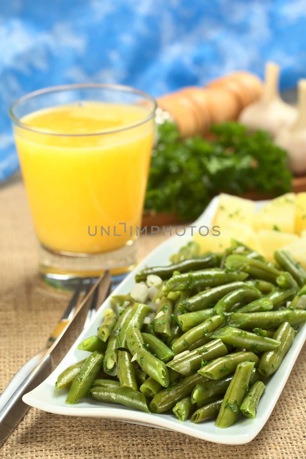 Cooked green beans with onion and parsley with cooked potato, orange juice and ingredients in the back (Selective Focus, Focus on the front of the beans)