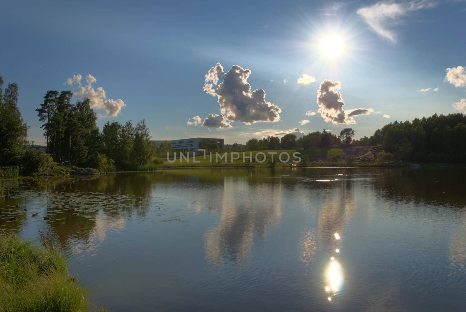 A still lake in the late summer with the Sun, clouds and a school on a hill side. The image is HDR tonemapped from 3 exposures with detail enhancement.
