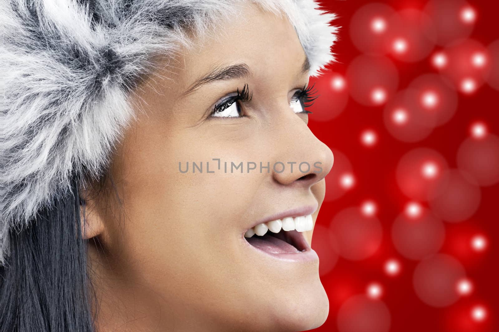 Christmas magic concept: beautiful young woman wearing grey faux fur hat, profile portrait looking up smiling and laughing over bright red background with bokeh light effects.
