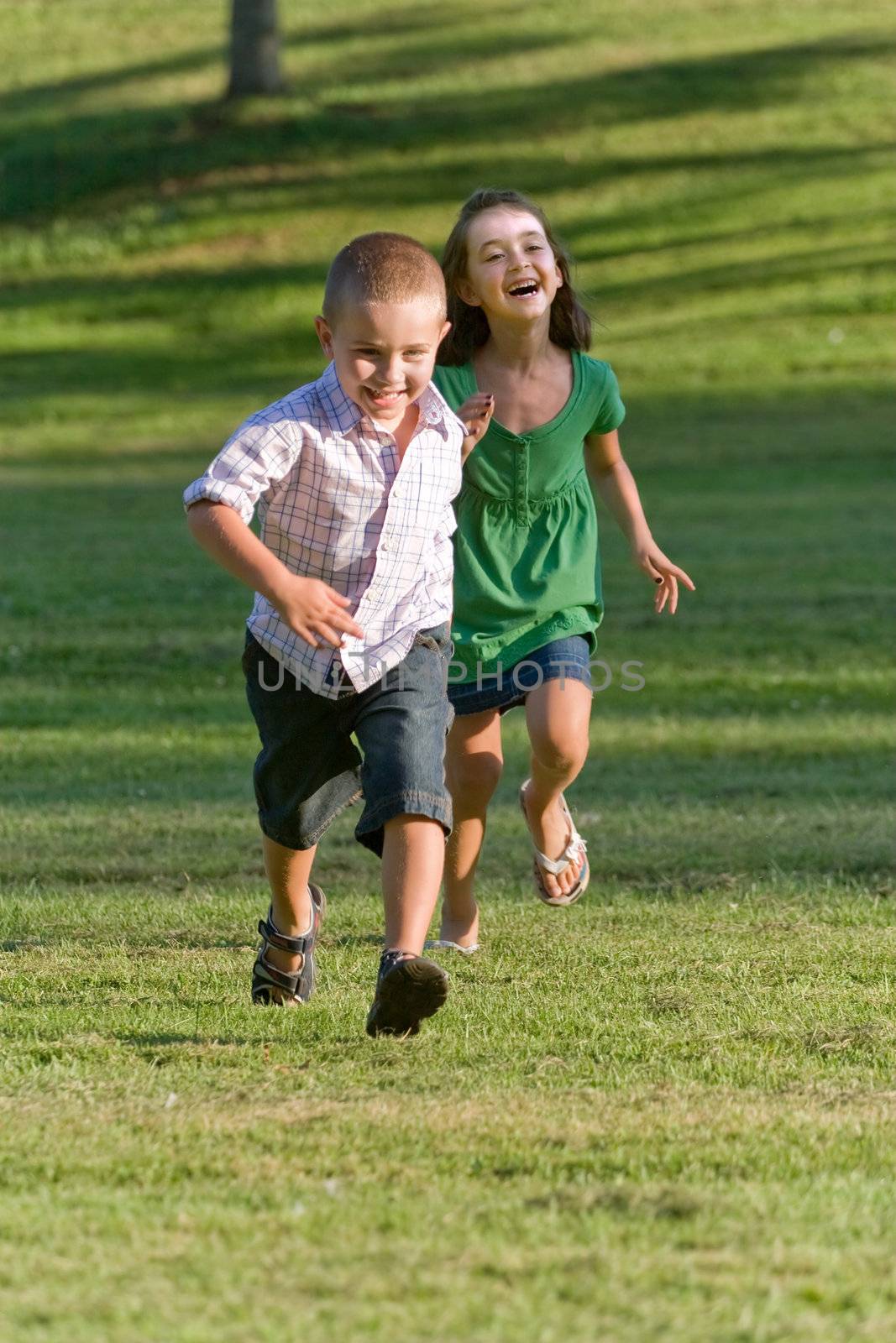 A young brother and sister running through a green grassy field with smiles on their faces.