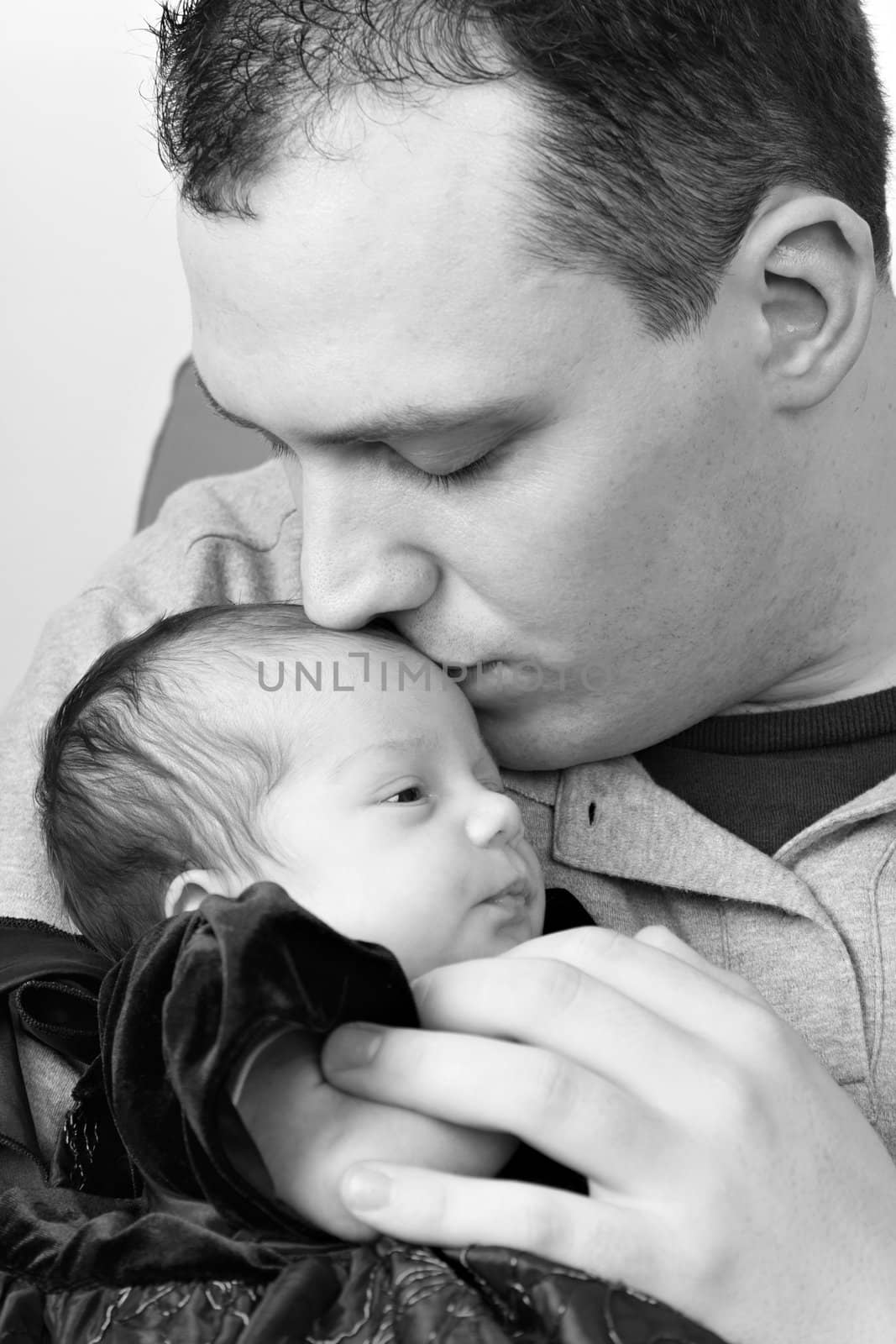 A newborn baby girl being by her dad as he kisses her head in black and white.