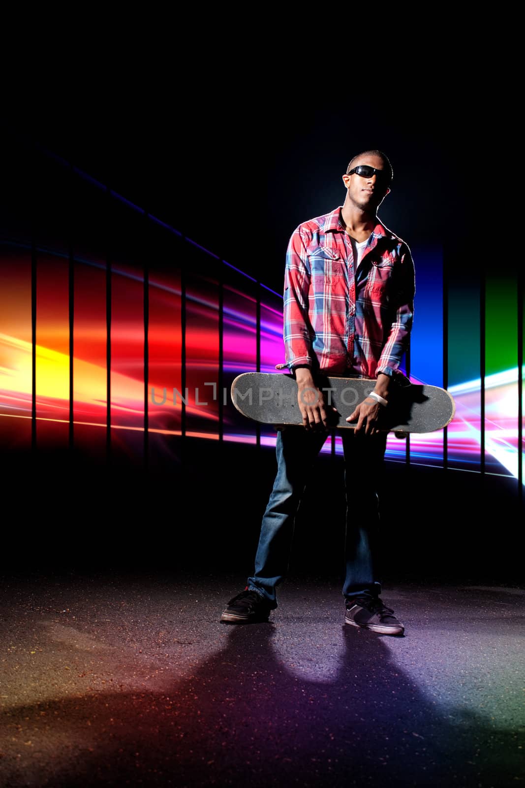 An African American skateboarder  holding his skateboard deck under dramatic lighting with a creative rainbow color schemed glowing spectrum in the background.
