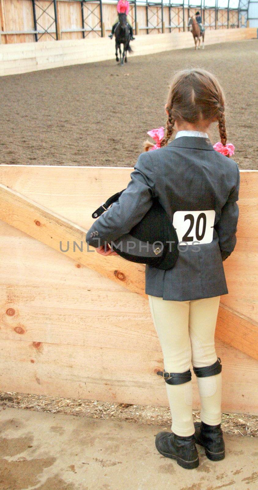a young female equestrian with riding helmet under her arm, patiently waits to be called to compete in the show ring