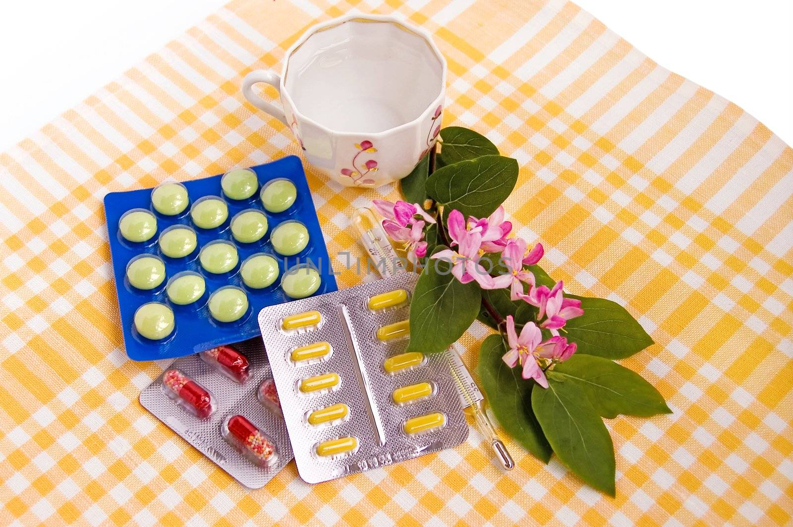 A set of tablets and capsules with a thermometer, the water in a porcelain cup and a branch with pink flowers and green leaves on a checkered yellow linen napkin