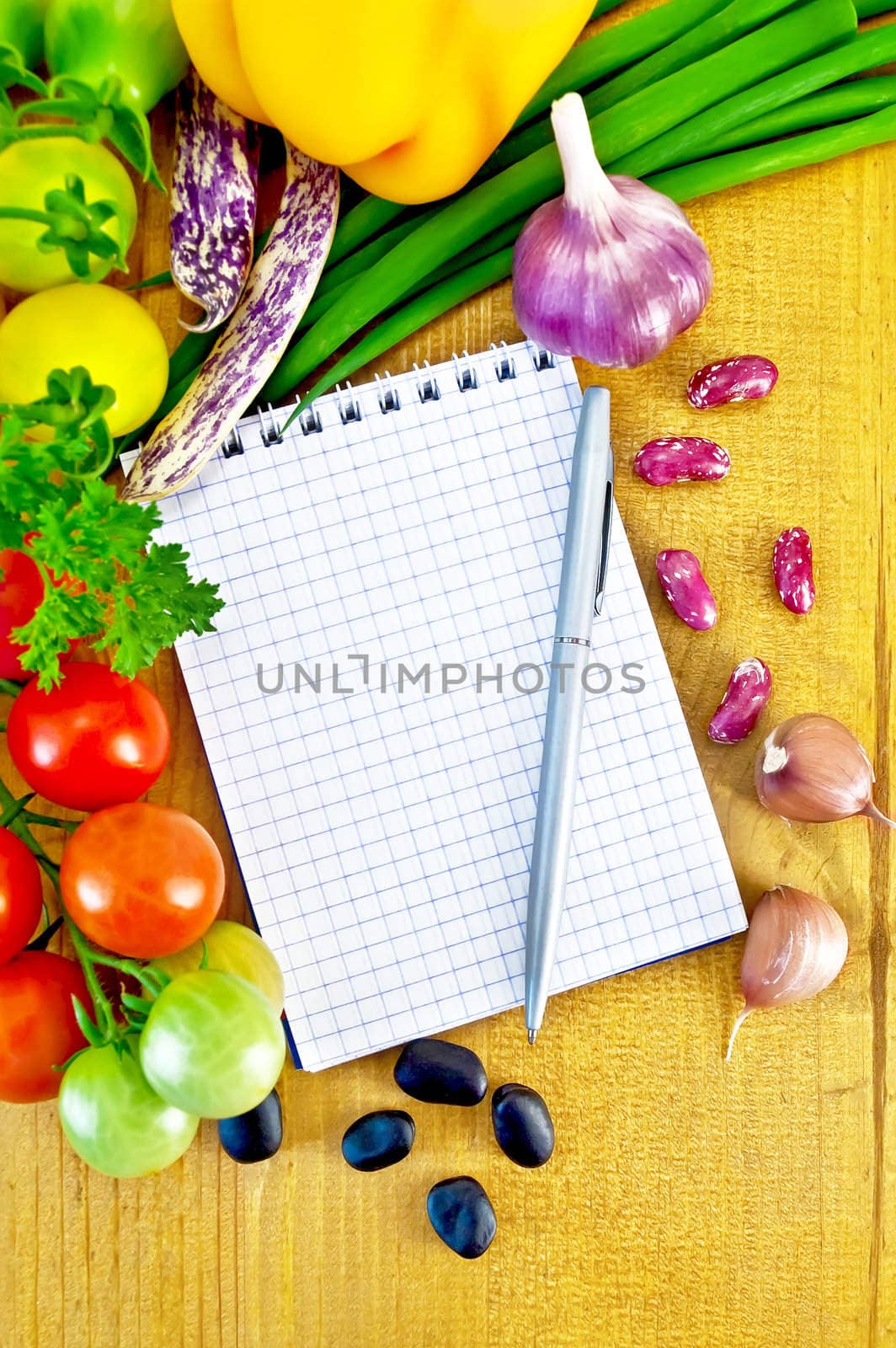 Notebook, silver metallic pen, small twigs of tomatoes, yellow bell pepper, green onions, parsley, whole and two cloves garlic, black beans and kidney beans on a wooden board