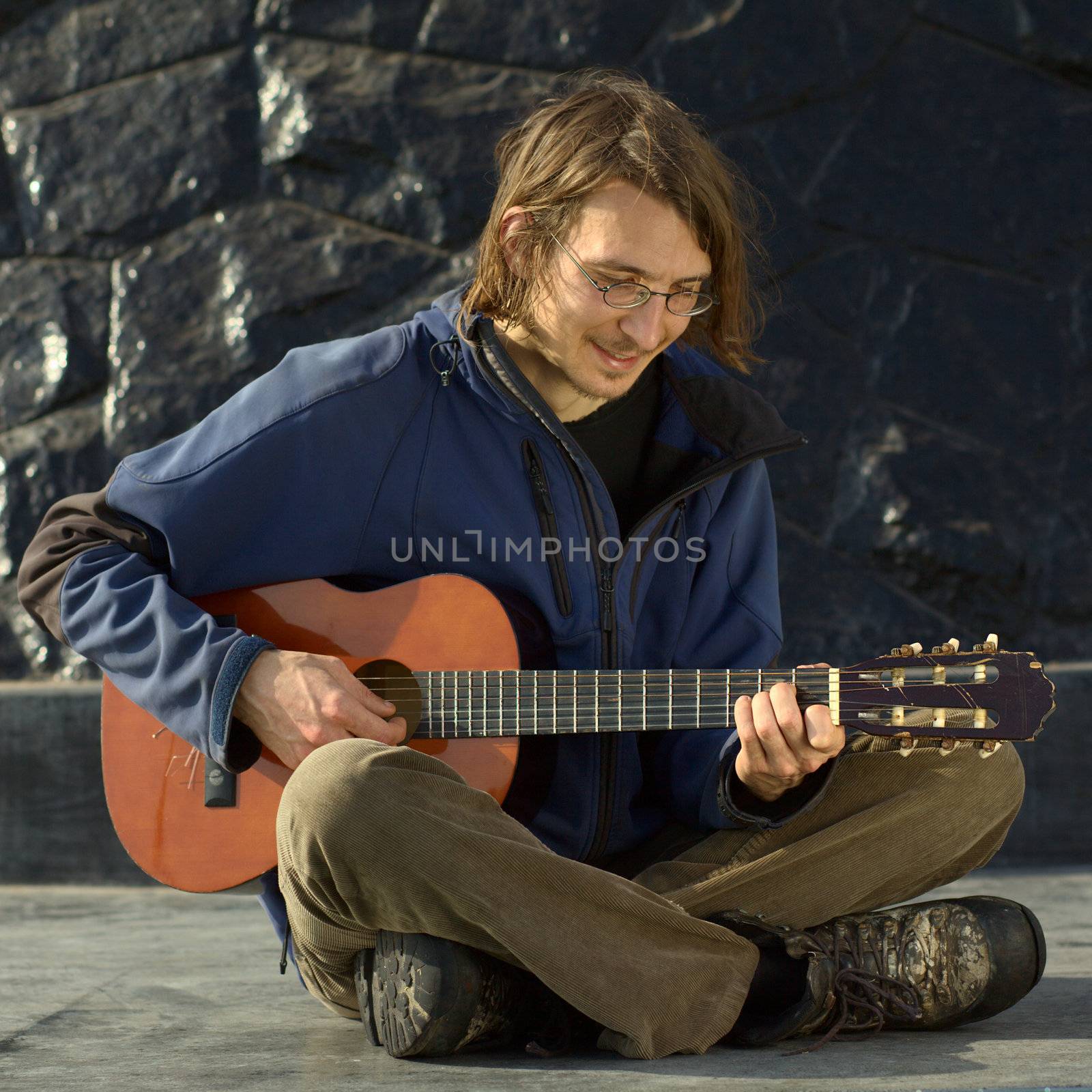 Young smiling Caucasian man playing the guitar sitting cross-legged outdoors on the ground 