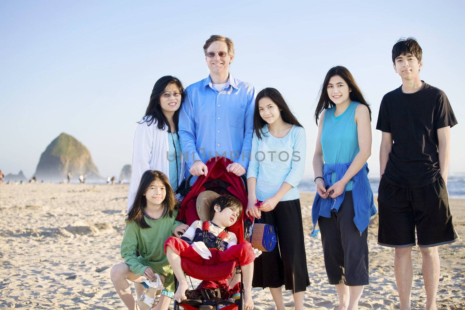 Large family of seven standing on the beach by the ocean. Boy in stroller is disabled with cerebral palsy.