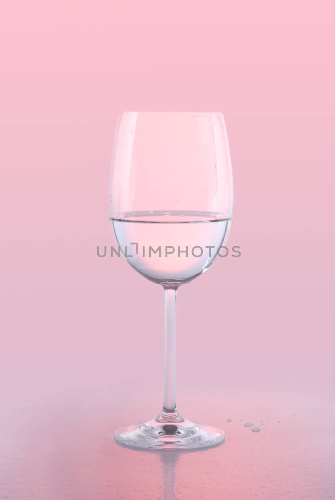 A crystal white wine glass holding water against a pink background with reflection and water drops.