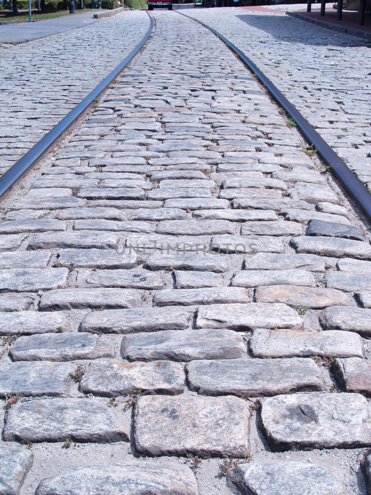 Train tracks embedded in down the middle of an old cobblestone road. The tracks curve into the distance in this perspective view.