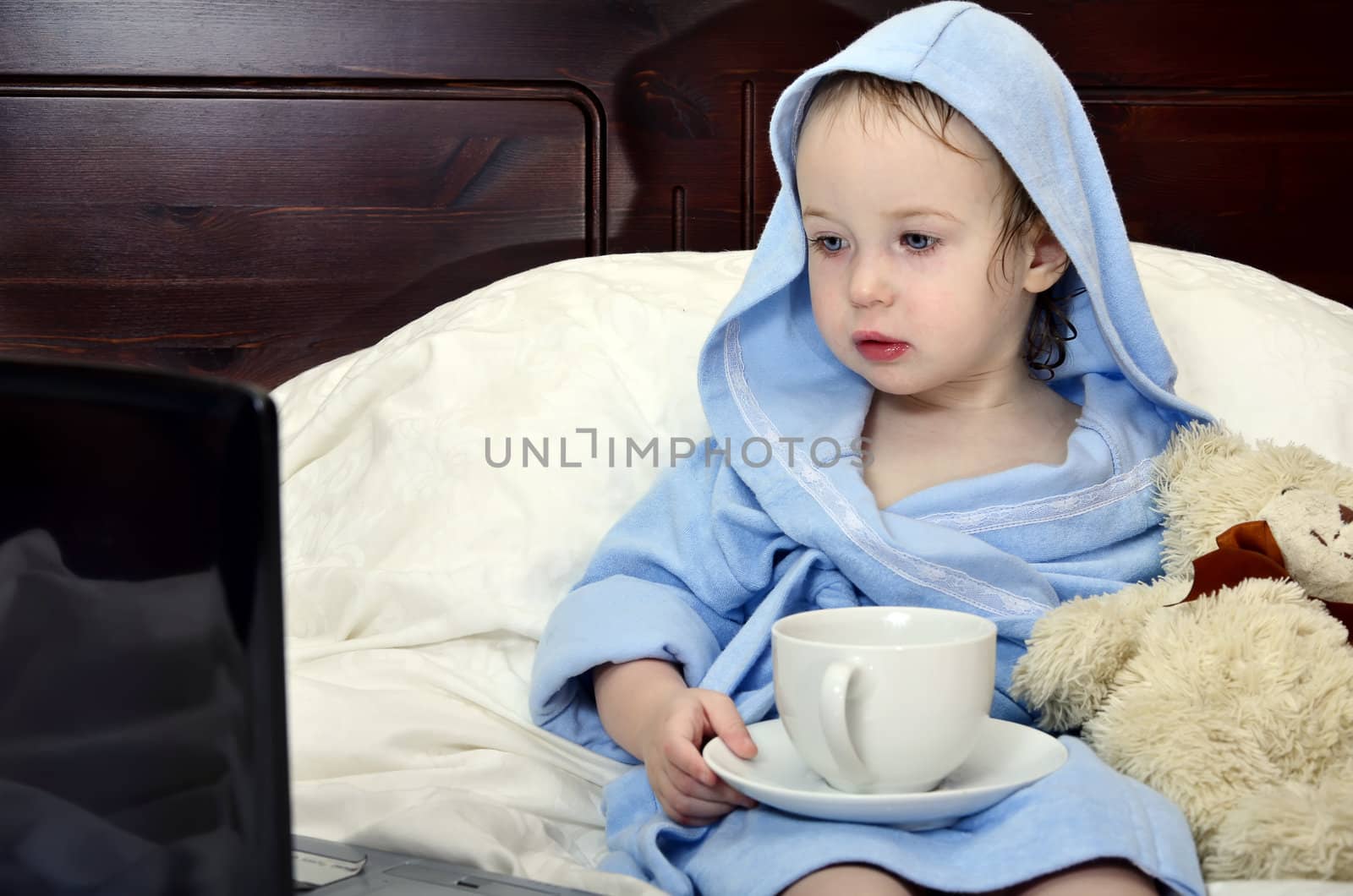 little girl in a bathrobe relaxing on the bed after a shower with cup of tea and laptop