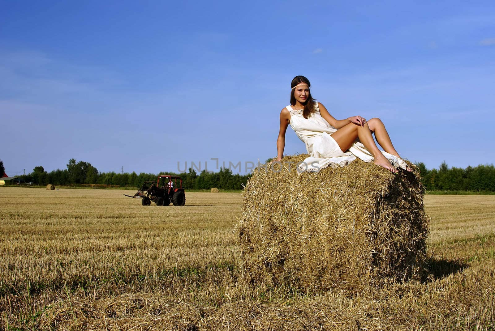 girl in a rural clothing sitting on the haystack, tractor on the background. space for text
