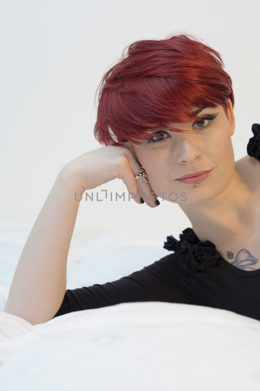 Portrait of a girl with red hair, piercing on lower lip, and tattoos on chest and shoulders, on quilt and serious expression.