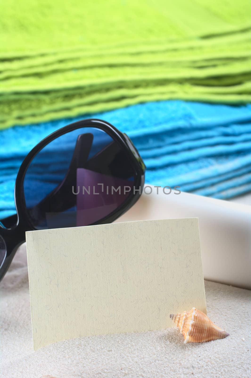Blank card on sand with a shell in front, and sunglasses, suncream and colorful beach towels in the back (Selective Focus, Focus on the card and the shell)