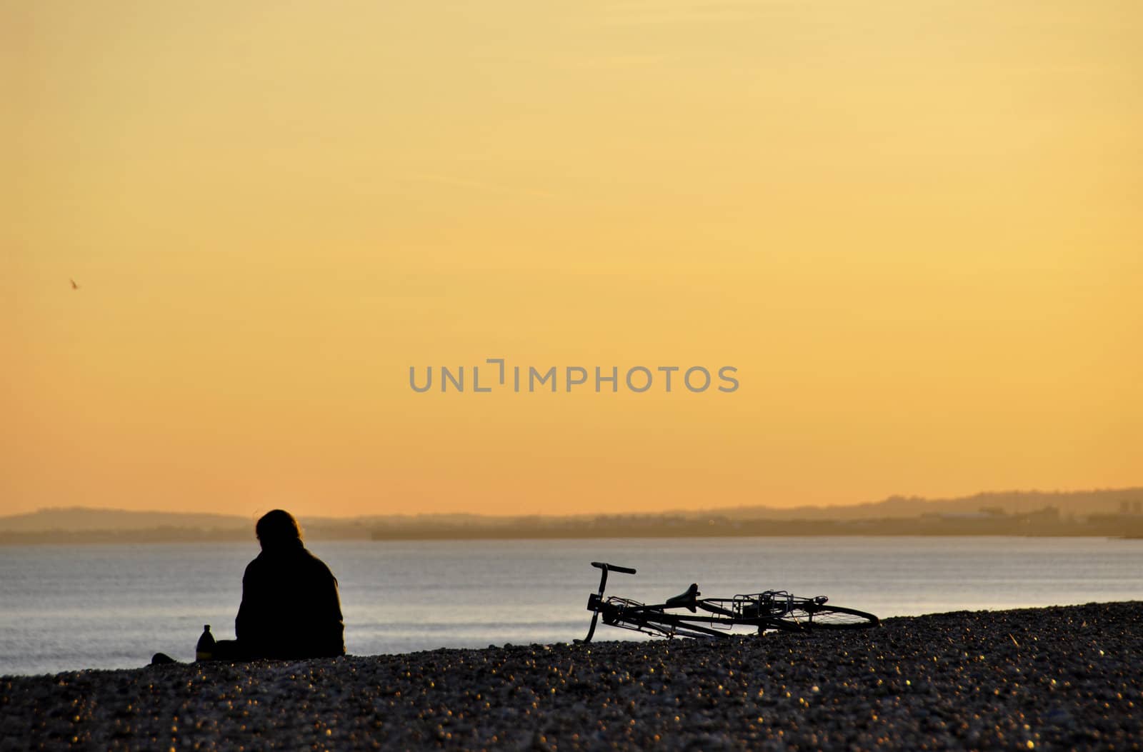 A sitting man and his bicycle on a pebble beach at sunset