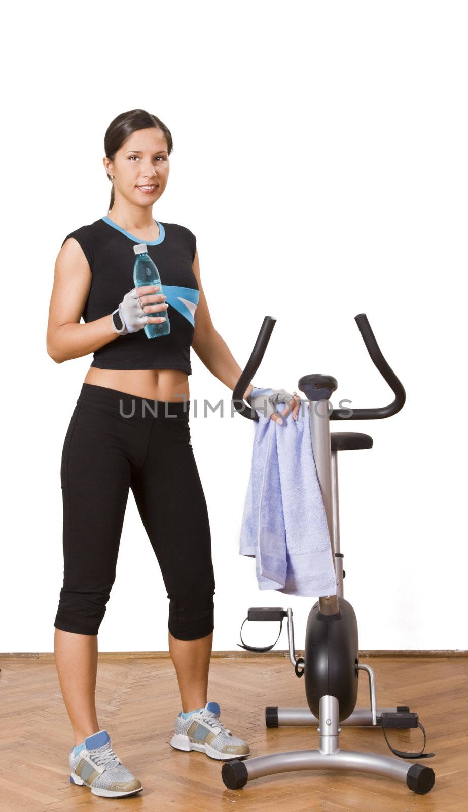 A young woman standing near a fitness bike with a glass of water in her hand.