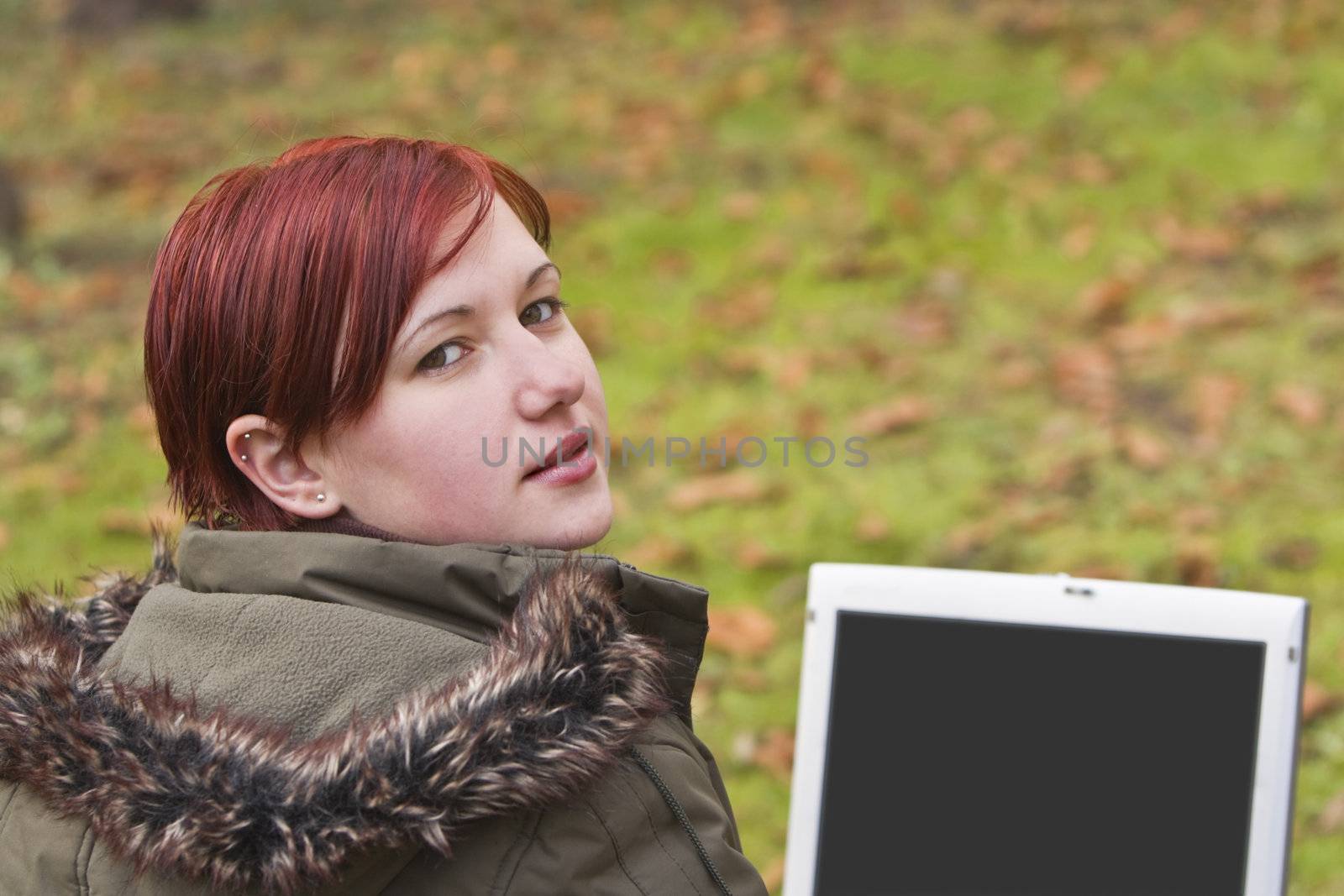 Redhead girl with computer in an autumn park looking up-back over the shoulder.