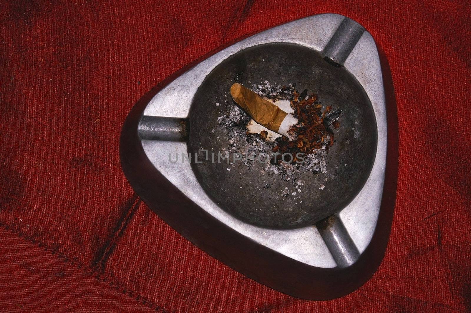 dirty metal ash tray with single stubbing sigaret in it on a red cloth   by karinclaus