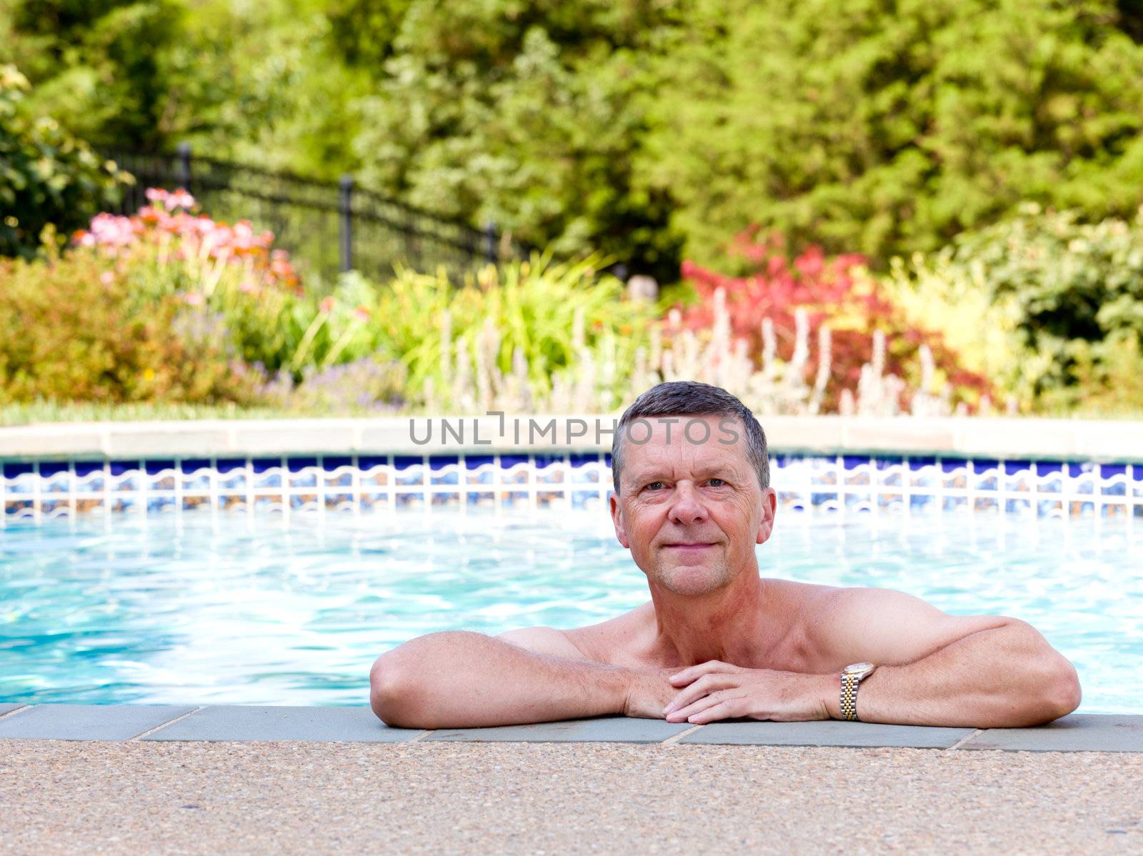 Senior male relaxing by the side of a modern swimming pool in back yard garden and facing the camera with smile