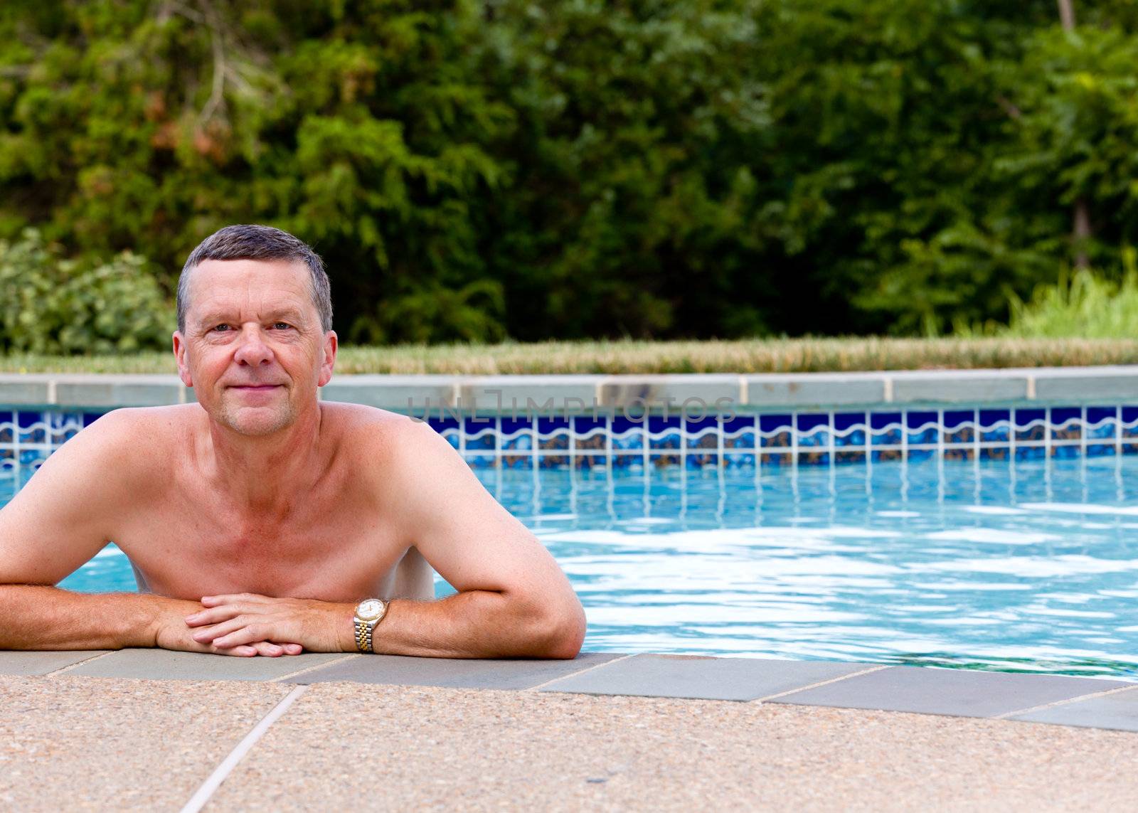 Senior male relaxing by the side of a modern swimming pool in back yard garden and facing the camera with smile