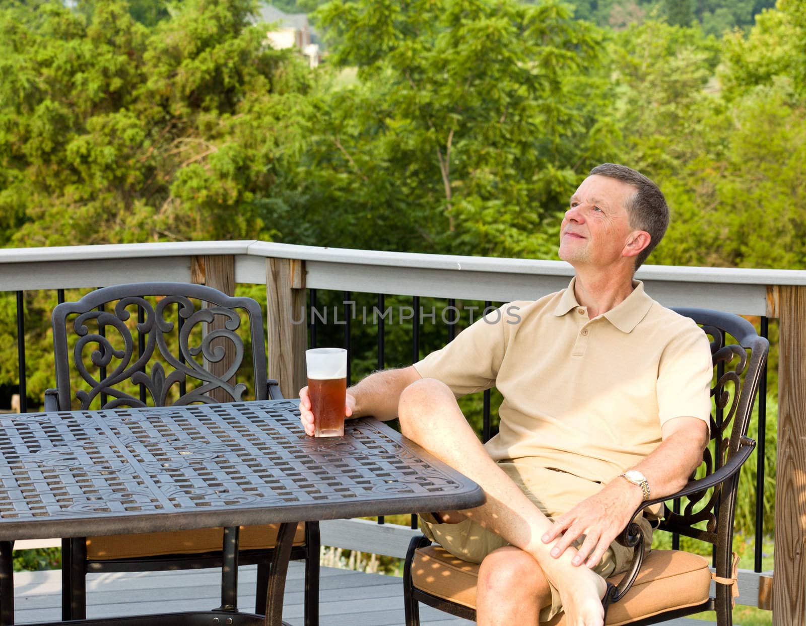 Middle aged man sitting on cast aluminium garden table on deck and drinking a glass of beer in back yard