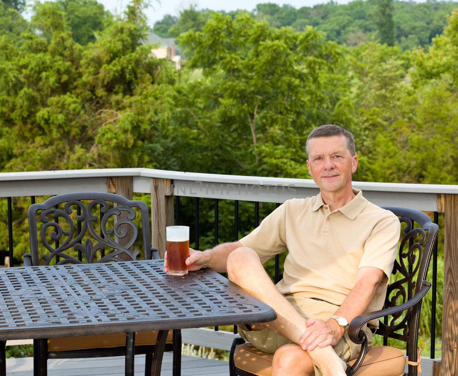 Middle aged man sitting on cast aluminium garden table on deck and drinking a glass of beer in back yard