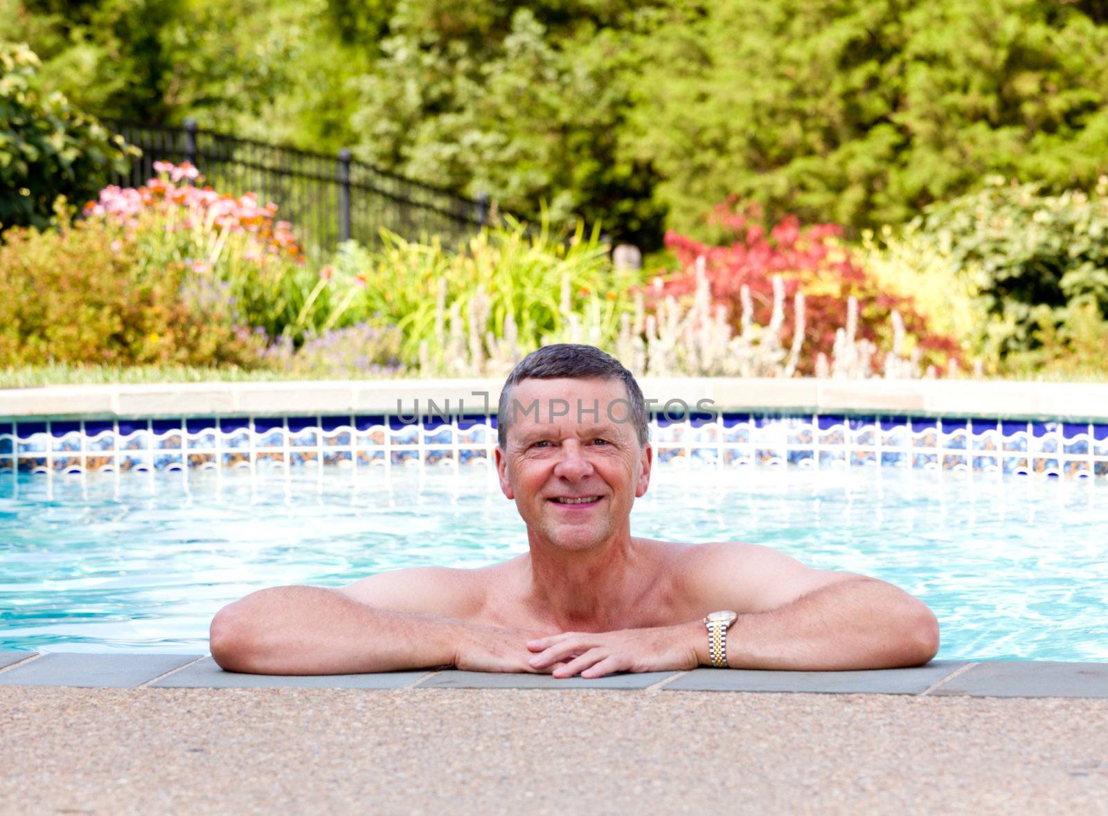 Senior male relaxing by the side of a modern swimming pool in back yard garden and facing the camera with smile