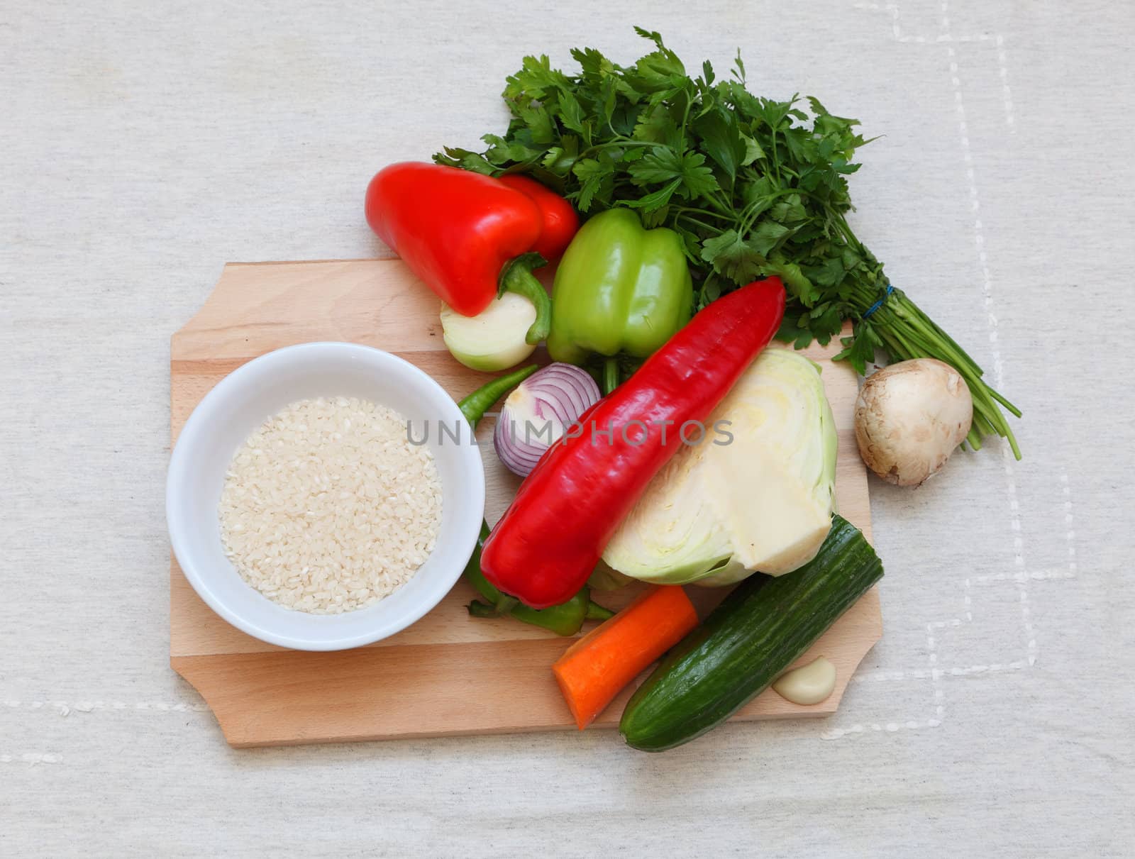 Upper view of various vegetables and a bowl of rice on a chopping board on a table.