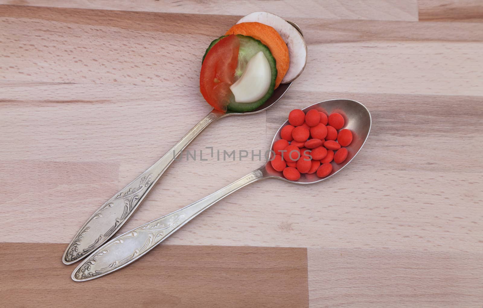 A spoon with fresh vegetables and one with red tablets on a wooden board.