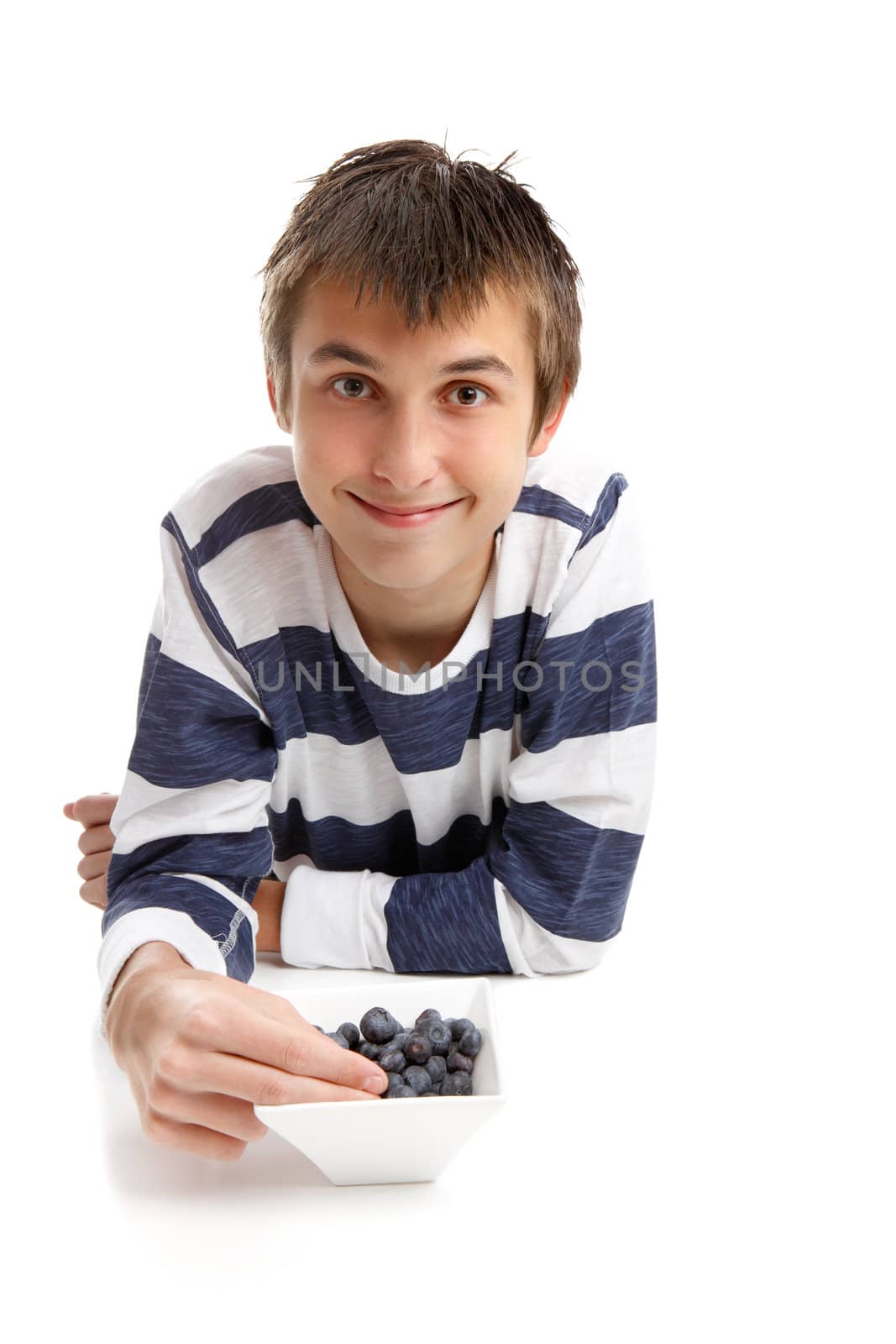 A young boy wearing a striped shirt is relaxing, eating from a bowl of fresh healthy blueberries.  White background, space for copy.