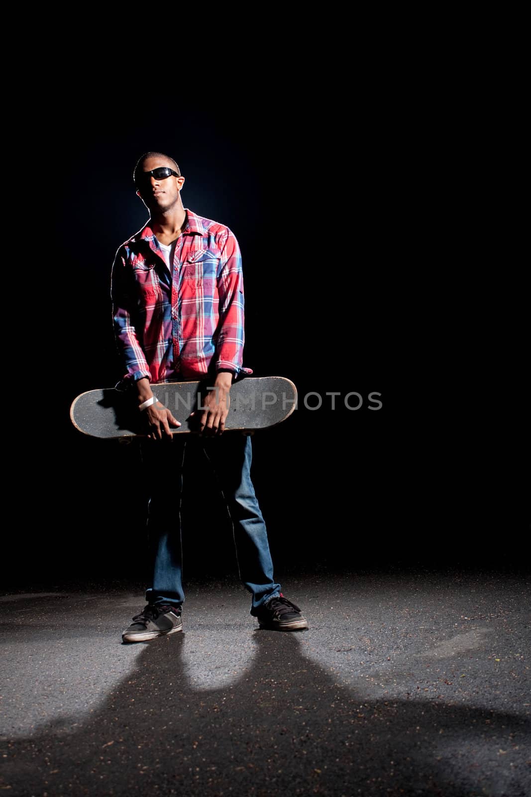 African American skateboarder wearing sunglasses holding his skateboard under dramatic lighting with dramatic shadows.  