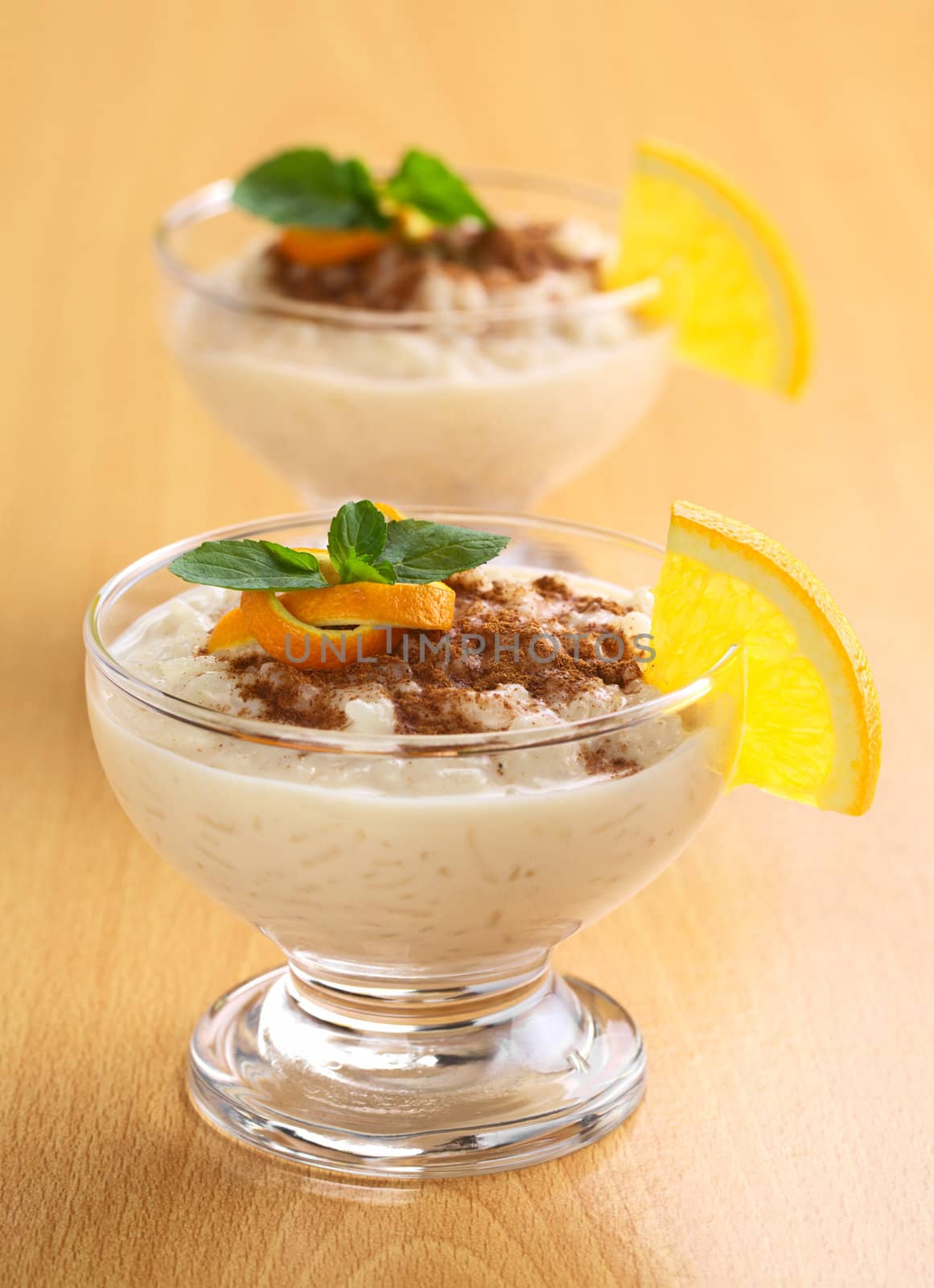 Delicious homemade rice pudding with cinnamon garnished with orange peel and mint leaf and a slice of orange on the rim of the bowl (Selective Focus, Focus on the orange peel and the mint leaf as well as the orange slice on the rim of the first dessert)