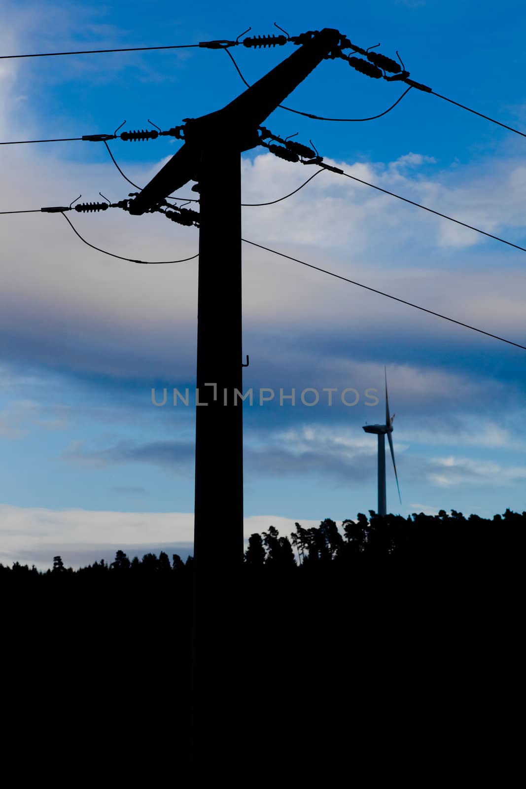 Silhouettes of large wind turbine on the horizon rising high above forest tree tops with high-voltage transmission line in foreground.