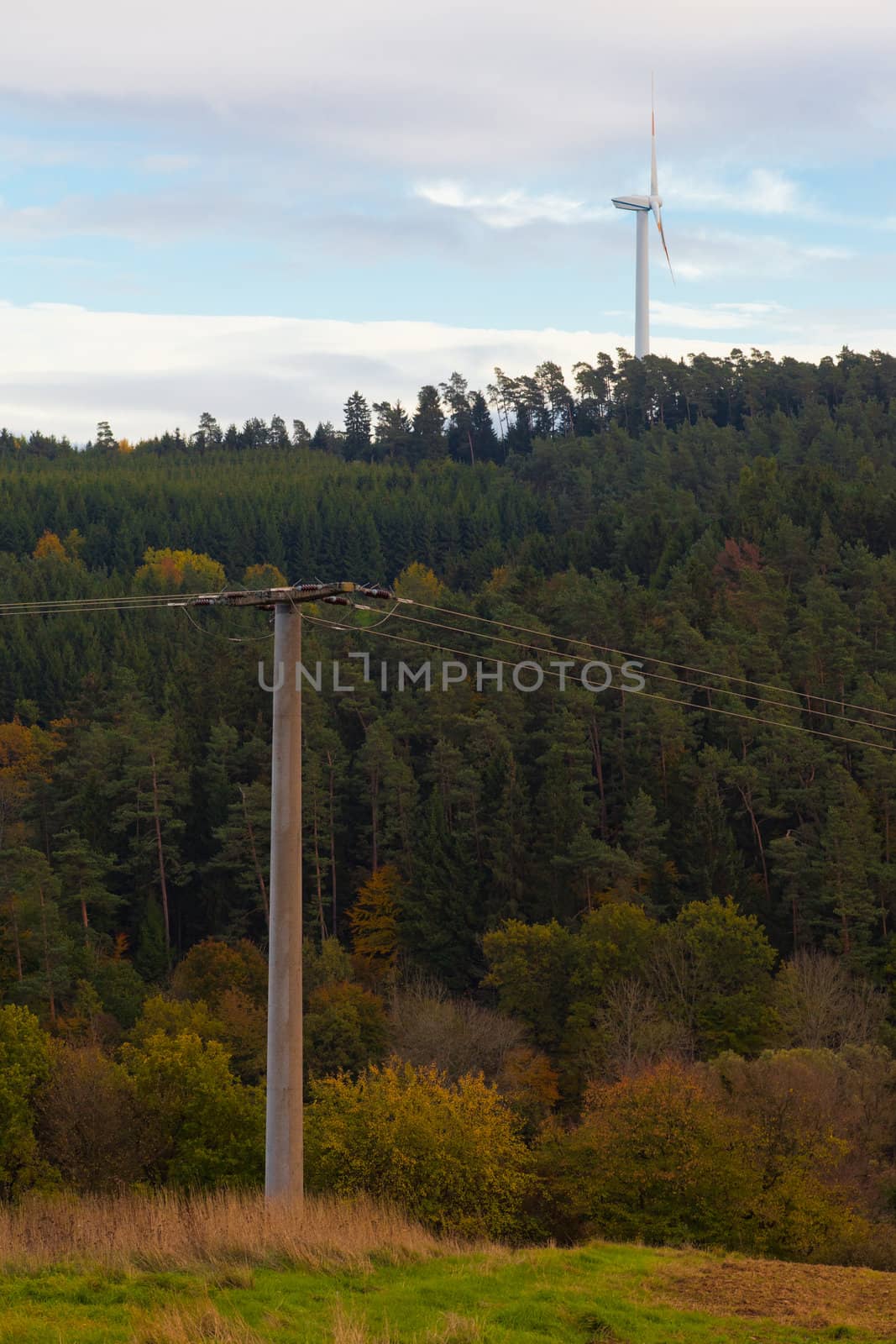 Large wind turbine on the horizon rising high above forest tree tops with high-voltage transmission line in foreground.
