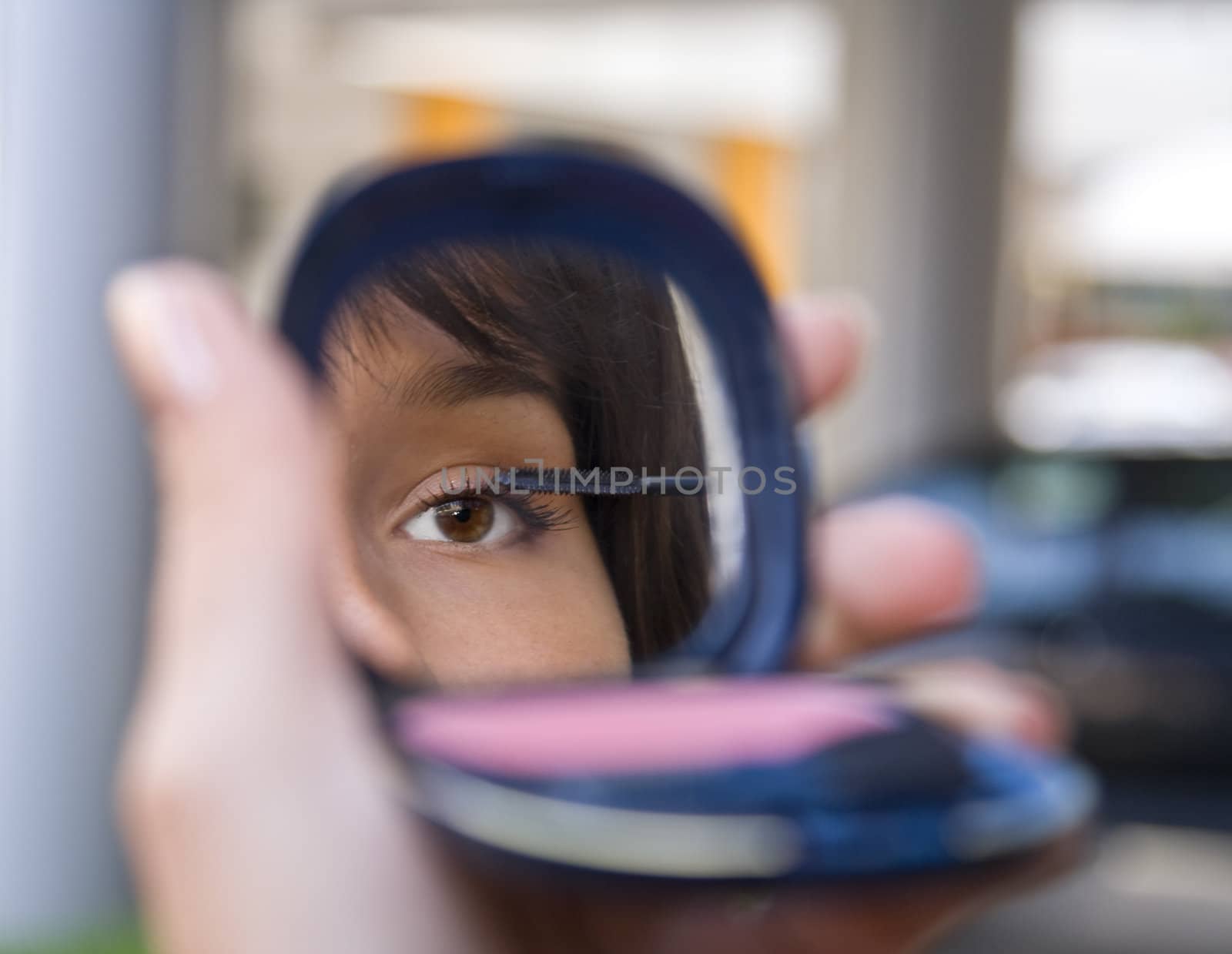 Mirror image of a woman's eye while she is applying mascara against a urban blured background.