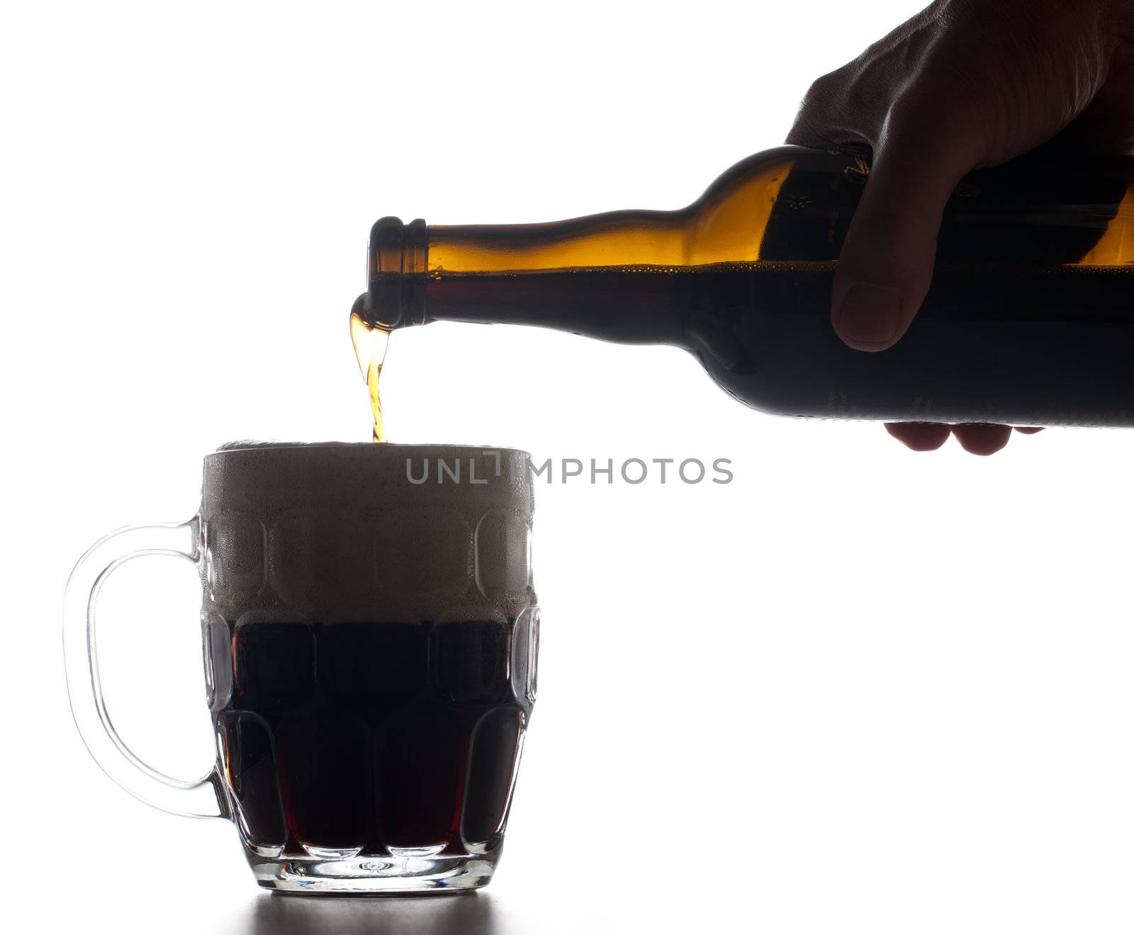 Backlit isolated beer tankard with dark ale being poured from brown bottle and creating a large head of froth on the liquid